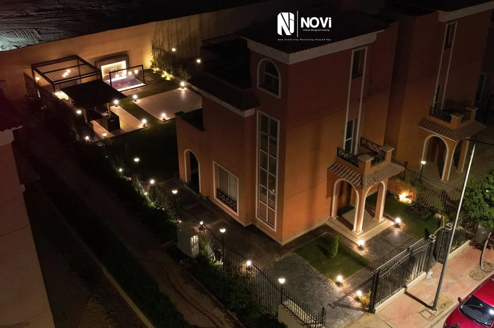 The villa garden seen from above at dusk: lit pergolas, a lawn and a lantern-lined path beside the villa